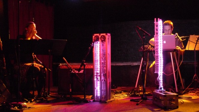 Troy Rogers performs behind the illuminated pink columns of the robotic musical instruments AMI and CARI, which form Expressive Machines Musical Instruments' (EMMI) MARIE, with Dana Jessen of the EAR Duo at the Southern in Charlottesville, VA