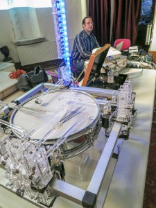 Troy Rogers performs music for robots at Sacred Heart Music Center in Duluth, MN. A robotic drum (MADI: Multi-mallet Automatic Drumming Instrument) is shown in the foreground, with robotic clarinet (CARI: Cylindrical Aerophone Robotic Instument) and vocal robot (Stemmetje) further back. Troy Rogers sits at his computer controlling the robots.